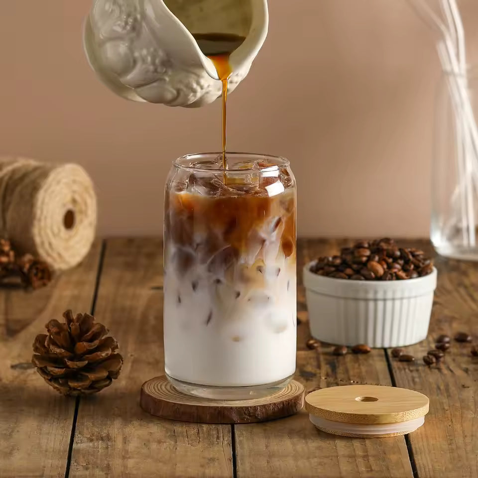 Pouring coffee into a glass of iced coffee and milk on a wooden table with coffee beans and a pine cone.